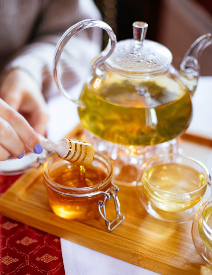 Elegant glass teapot with herbal tea, honey drizzle, and cups on a wooden tray for a perfect tea time setting.